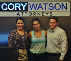 three people stand under a Cory Watson Attorneys sign