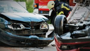 A blue car has a destroyed front end with the bumper hanging off and a red car flipped on it's hood lays in front of it. Cory Watson Attorneys Personal Injury law firm works closely with auto accidents or car wreck cases.